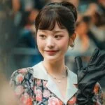 Женщина в цветочном платье и черных перчатках улыбается и машет рукой